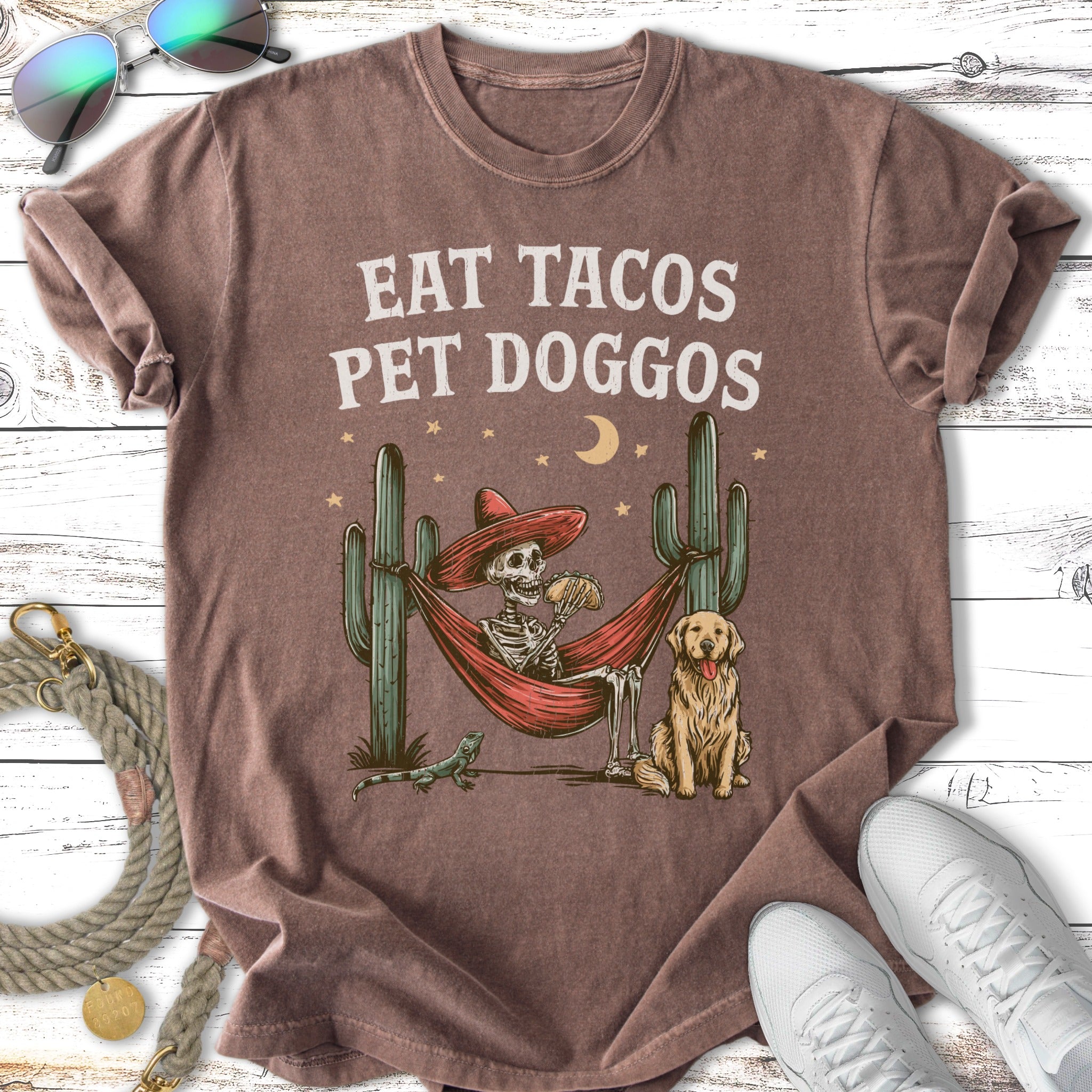 T-shirt with skeleton in hammock, dog, cactus, and text "EAT TACOS PET DOGGOS".