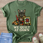 French bulldog wearing knight's helmet and cape, holding sewing tools, text "WE QUILT AT DAWN".