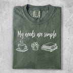 Green t-shirt with text "My needs are simple" and icons of coffee, paw print, and books.