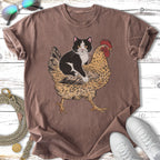 Tuxedo cat riding a chicken on a brown t-shirt.