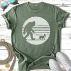 Green t-shirt with Bigfoot walking a small dog silhouette in a circle.
