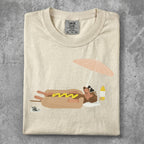 Cream t-shirt with cartoon hot dog in bun relaxing on beach under umbrella.