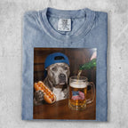 Dog wearing blue baseball cap holds hot dog next to beer with American flag flag.