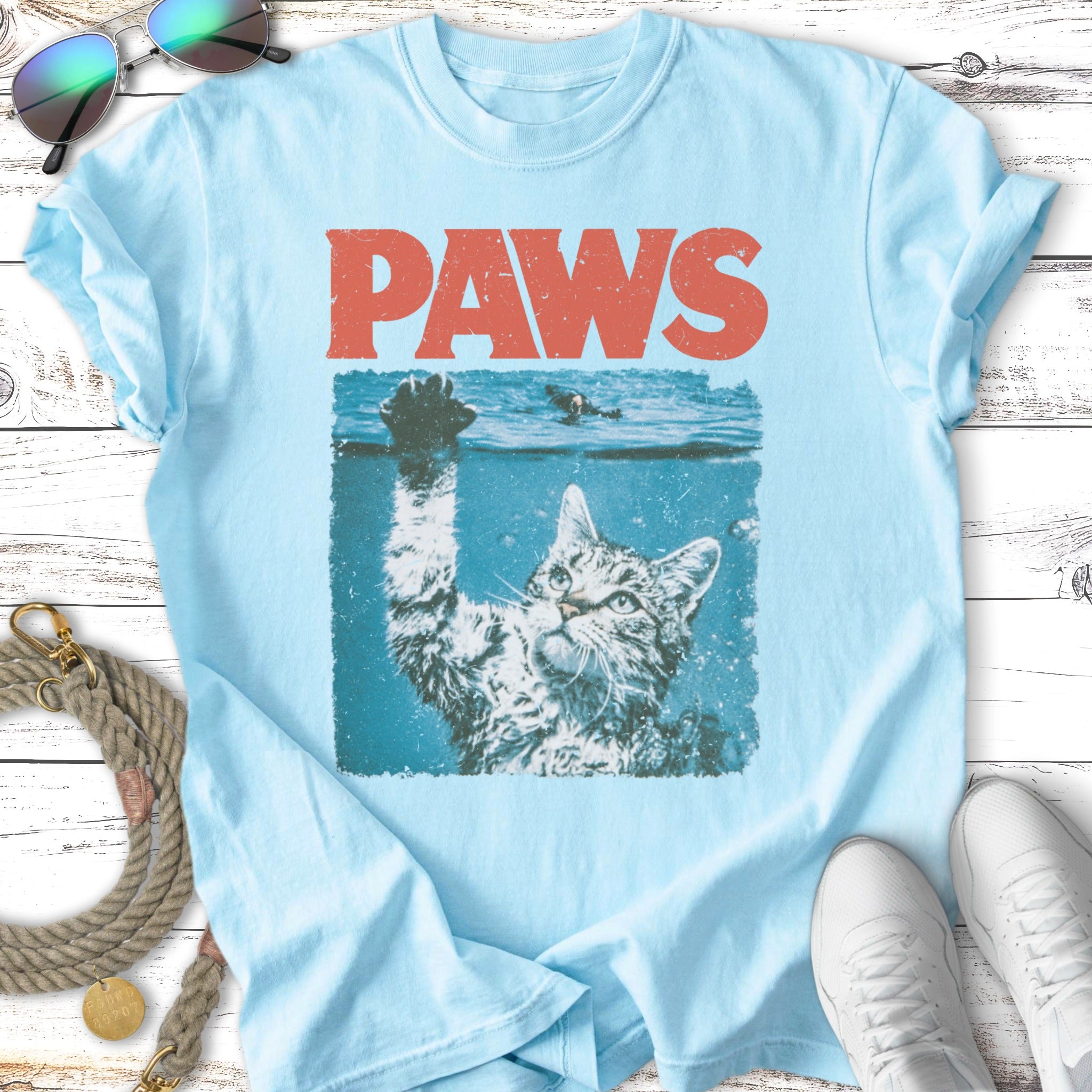 Light blue t-shirt with "PAWS" text above a distressed graphic of a cat's head emerging from water.