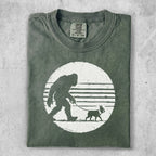 T-shirt graphic of Bigfoot walking a dog on a leash, white design on green shirt.