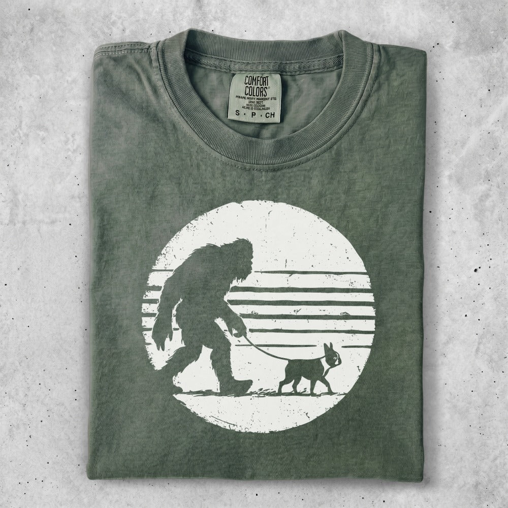 T-shirt graphic of Bigfoot walking a dog on a leash, white design on green shirt.
