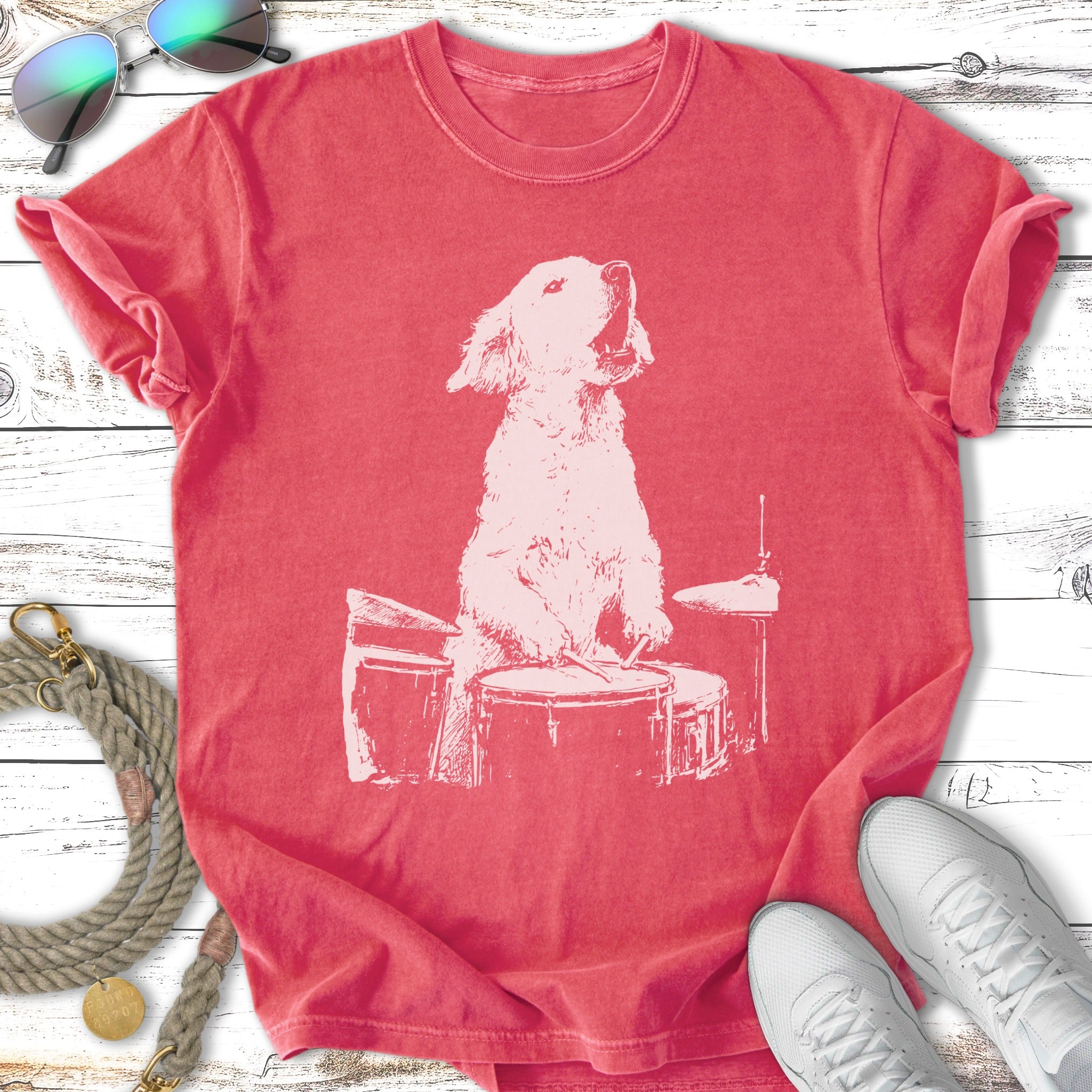 White graphic of a golden retriever playing a drum set on a coral t-shirt.