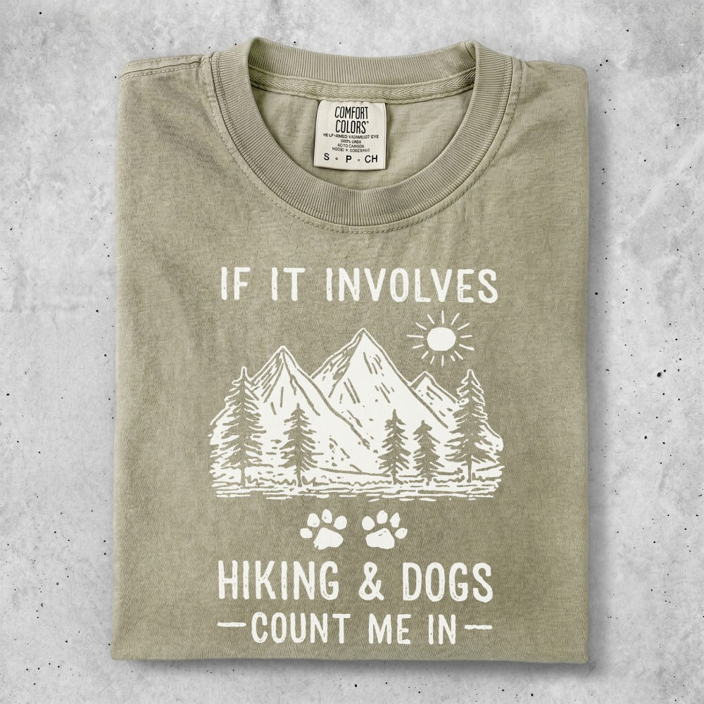 Graphic tee with mountains, trees, paw prints, and text "IF IT INVOLVES HIKING & DOGS - COUNT ME IN".