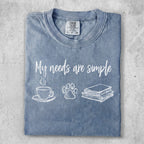 Blue t-shirt with "My needs are simple" text and coffee, paw print, and book icons.