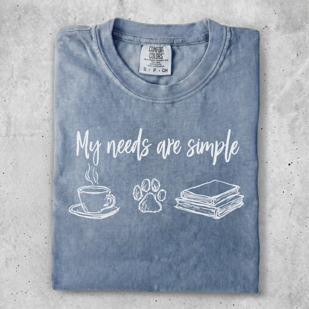 Blue t-shirt with "My needs are simple" text and coffee, paw print, and book icons.