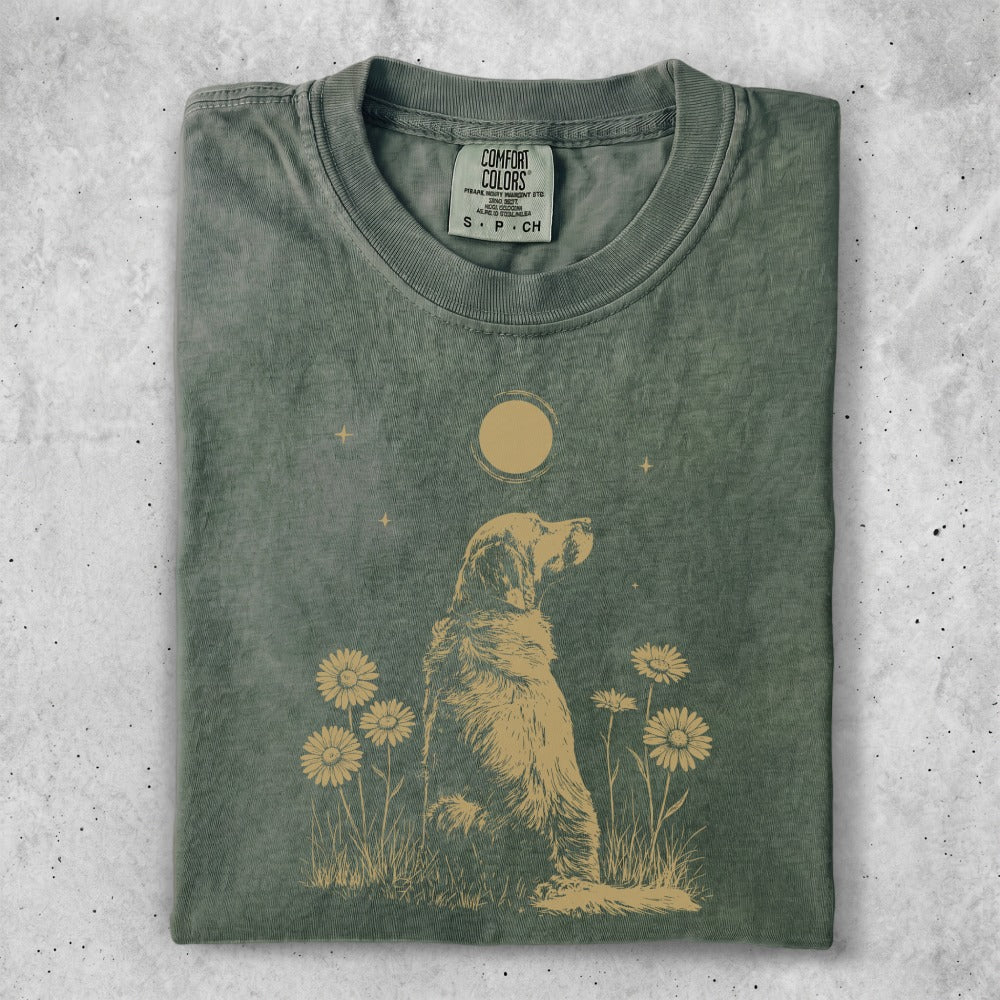 Golden retriever sitting in grass with sunflowers and moon design on green t-shirt.